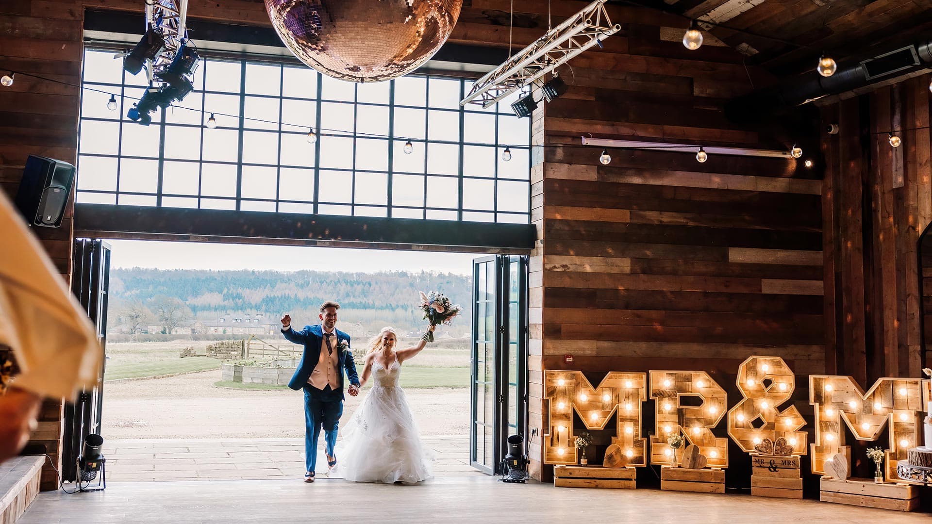 A bride and groom joyfully enter Wharfedale Grange, a rustic Yorkshire wedding venue with large windows. The bride holds a bouquet as marquee lights spell "MR & MRS" on the floor beneath a sparkling disco ball.