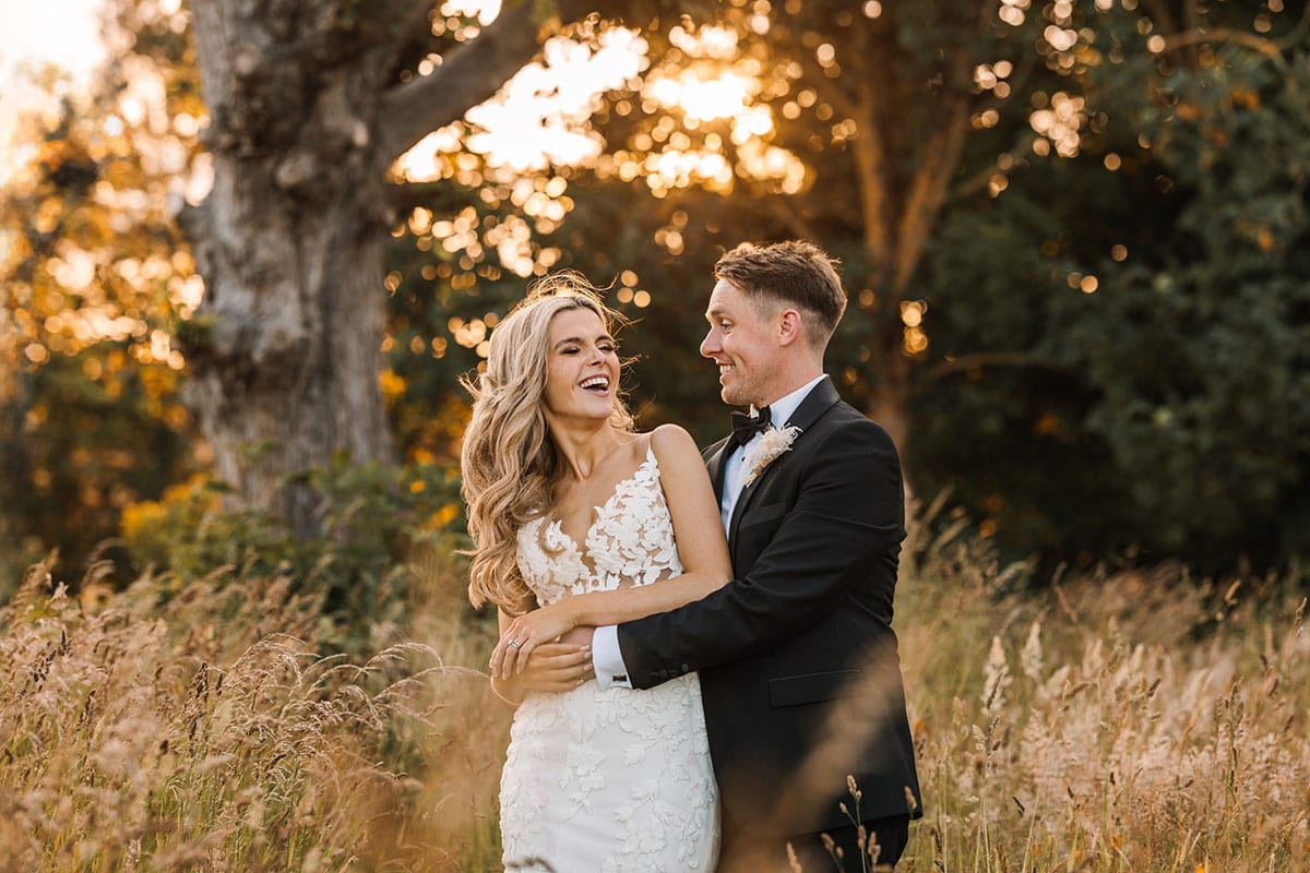 A bride in a white lace dress and a groom in a black tuxedo stand together in a Yorkshire field at sunset, smiling and embracing—capturing the magic of wedding photography.