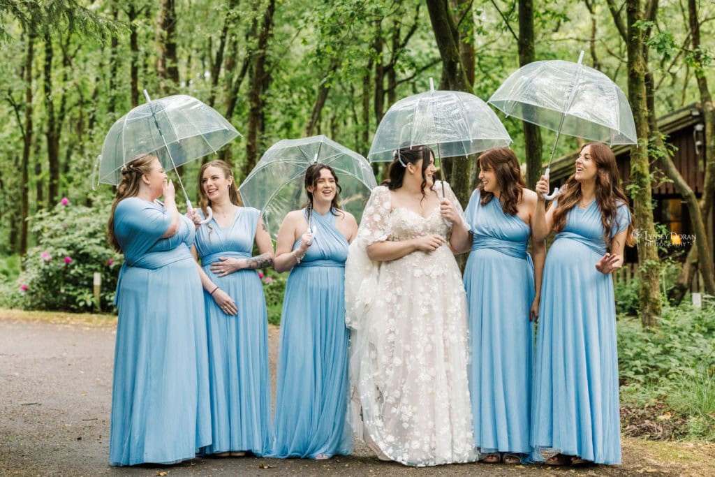 A bride in a white gown stands with five bridesmaids in matching light blue dresses, all holding clear umbrellas and laughing together—a perfect example of how to embrace rainy wedding day tips in a lush, green outdoor setting.