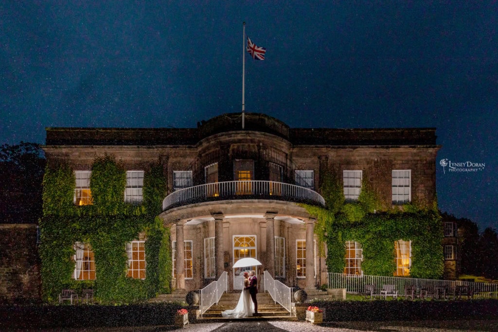 A bride and groom stand under a white umbrella in front of a grand, ivy-covered building at night, illuminated by warm lights. Rain falls around them, and a Union Jack flag flies atop the building.
