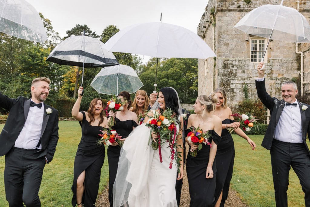 A bride walks outdoors with her bridesmaids and groomsmen, all smiling and holding clear umbrellas. The bridesmaids wear black dresses and hold colourful bouquets. The group is next to a stone building and lush greenery.