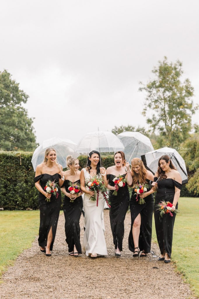 A bride walks outdoors with her bridesmaids and groomsmen, all smiling and holding clear umbrellas. The bridesmaids wear black dresses and hold colourful bouquets. The group is next to a stone building and lush greenery.