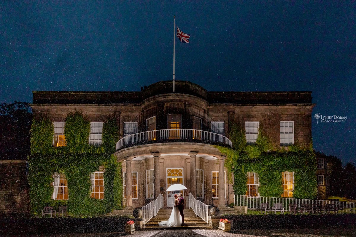 A bride and groom stand under a white umbrella in front of a grand, ivy-covered building at night, illuminated by warm lights. Rain falls around them, and a Union Jack flag flies atop the building.