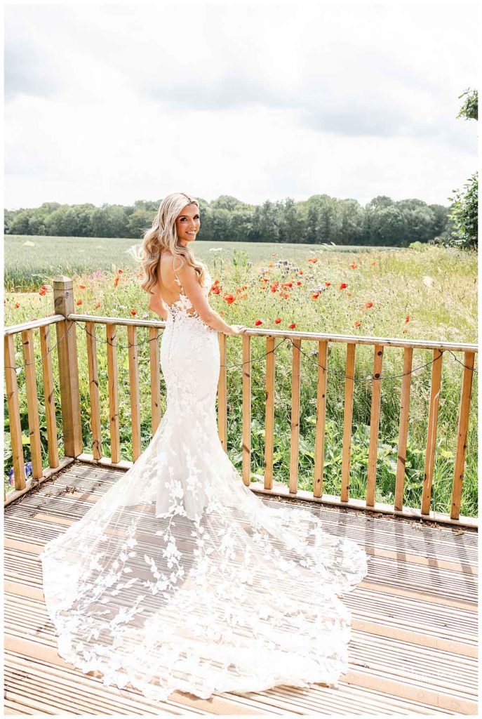 A bride in a fitted, floral lace wedding dress stands on a wooden decking, facing away and looking over her shoulder, with a field of wildflowers and trees in the background on a bright day.