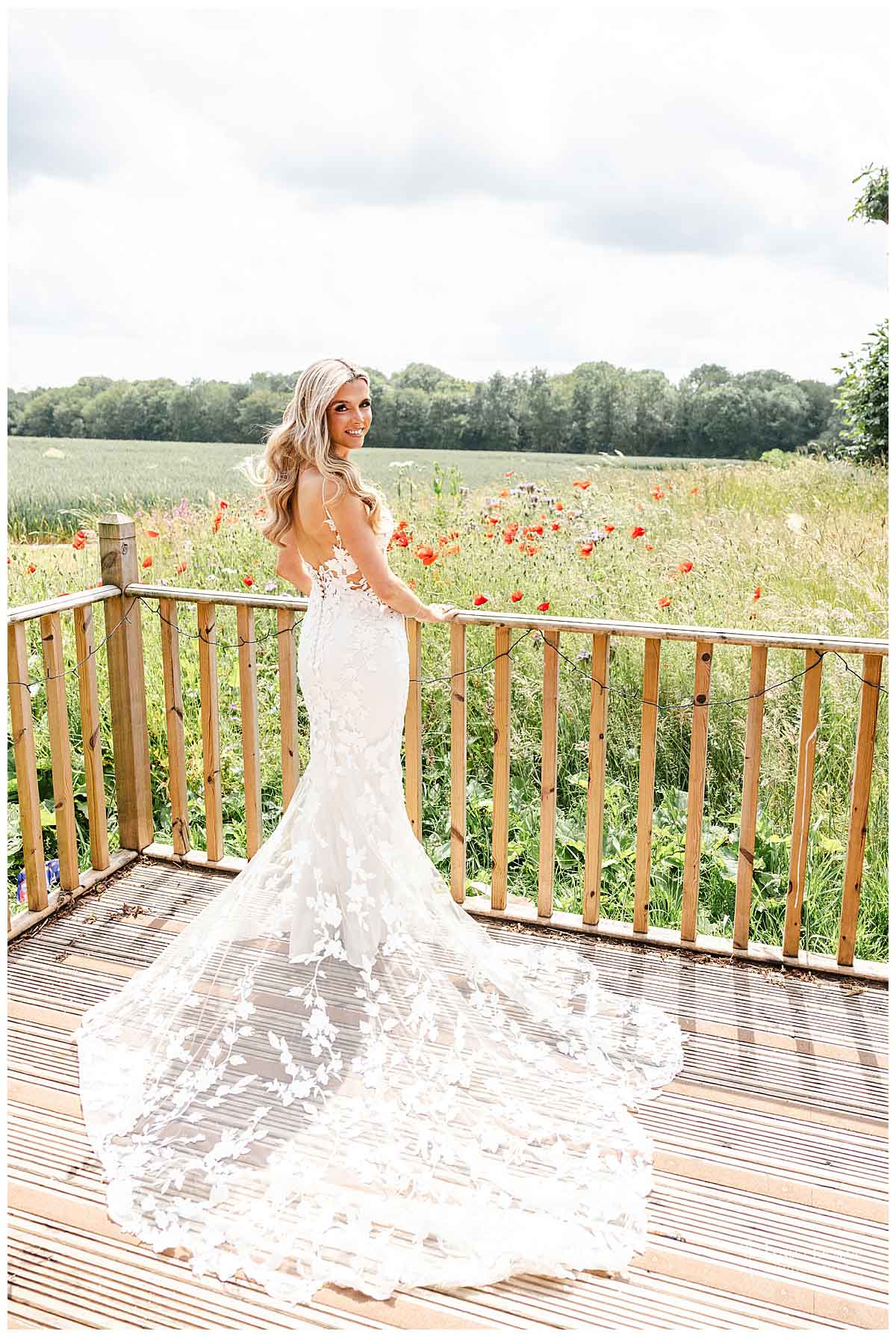 A bride in a fitted, floral lace wedding dress stands on a wooden decking, facing away and looking over her shoulder, with a field of wildflowers and trees in the background on a bright day.