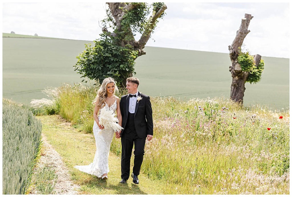 A bride in a white dress and a groom in a black tuxedo walk hand in hand on a grassy path through a field at their wedding venue in East Yorkshire, smiling at each other, with two uniquely shaped trees in the background. Taken by Hull wedding photographer