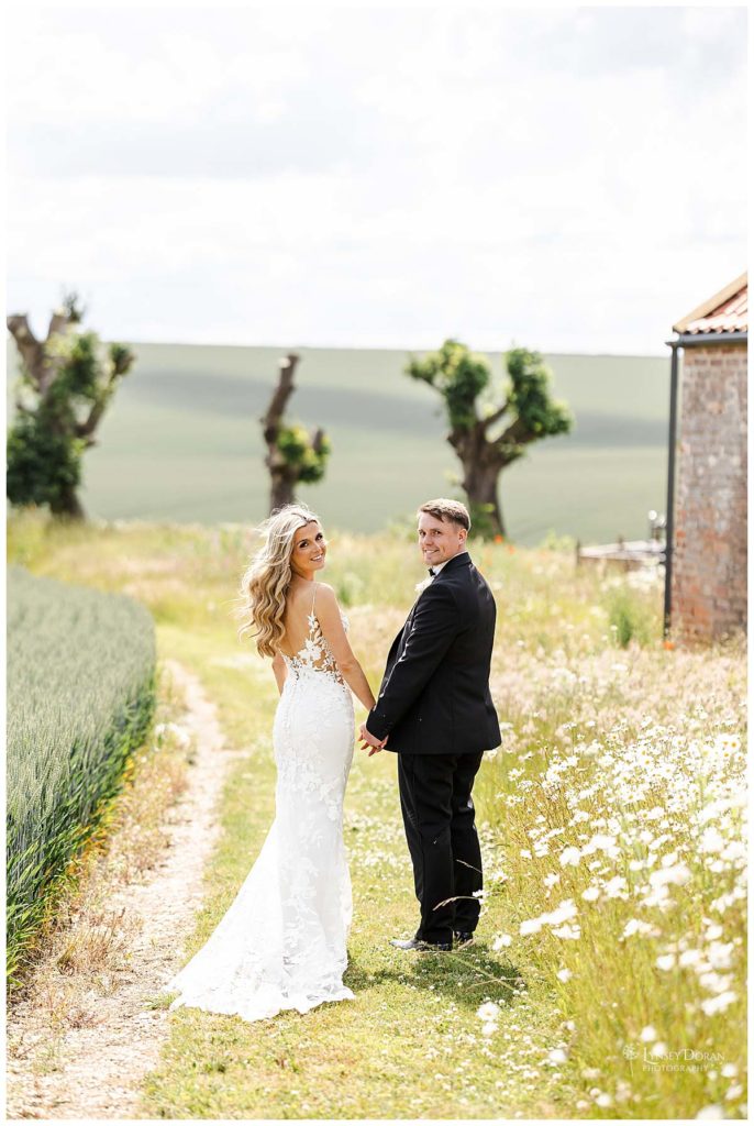 A bride and groom stand hand in hand on a rural path of their Hull wedding venue, surrounded by wildflowers and fields, smiling back at the camera. The bride wears a white lace dress; the groom is in a black suit. The wedding venue is Wold view farm East Yorkshire
