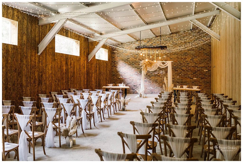 A rustic wedding ceremony room in a barn wedding venue in Yorkshire with rows of wooden chairs draped in white fabric, a brick wall backdrop, fairy lights overhead, and a decorated arch at the front.